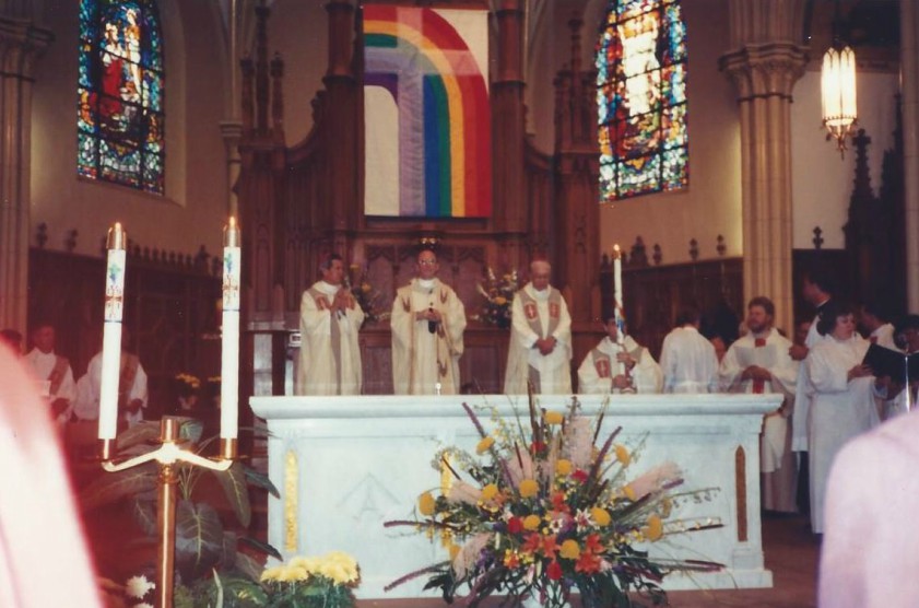 a groups of priests stand at the pulpit, behind them is a giant rainbow flag with a clear cross over the top of it