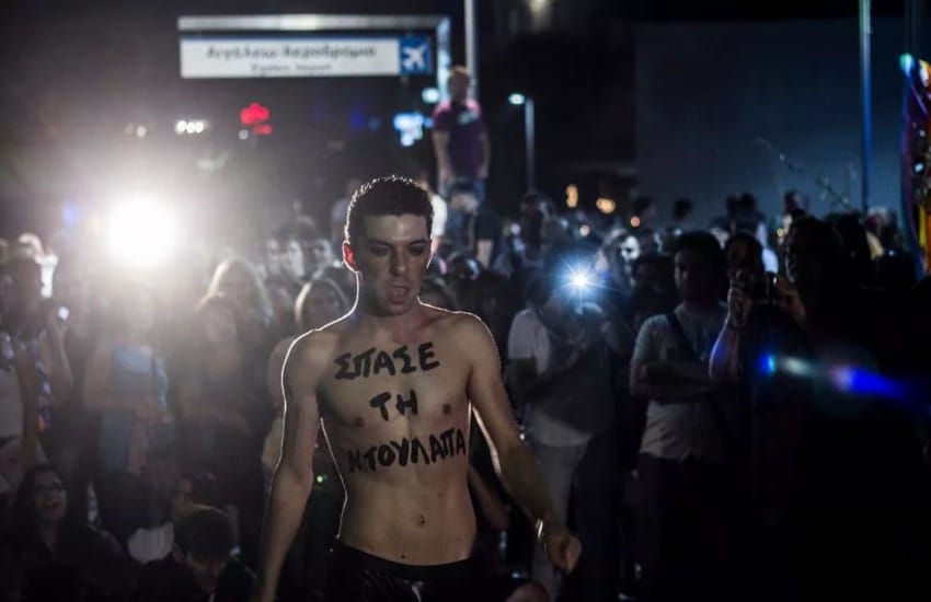a man standing topless at night surrounded by people. he has 'smash the closet' painted on his torso in greek