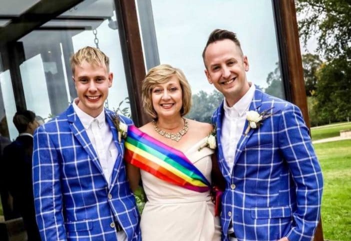 Vanessa Hall, her son, and his husband. The men are in blue suits and she is wearing the rainbow sash